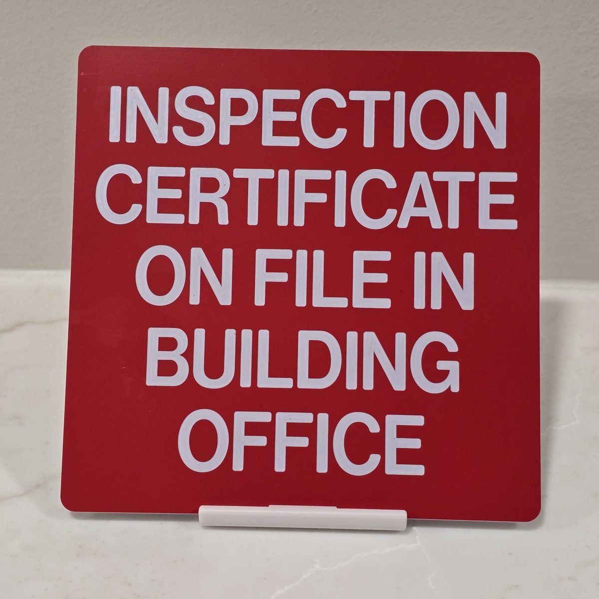 Red engraved sign reading Inspection Certificate On File In Building Office with white letters