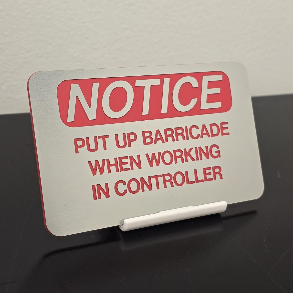 Brushed silver NOTICE sign reading Put Up Barricade When Working In Controller with red lettering